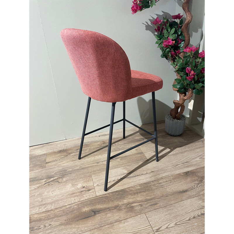 The Clio bar stool features a modern design with a curved red upholstered backrest and black metal legs, placed on a wooden floor beside a potted plant with pink flowers against a light gray wall.
