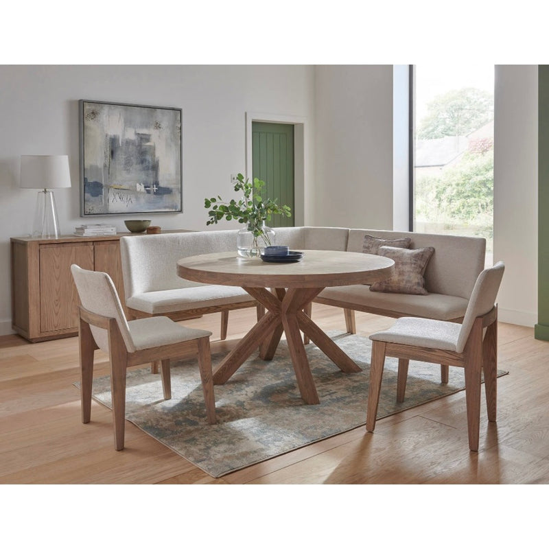 A modern round dining table with oak legs and a light beige fabric top, accompanied by four cream-colored dining chairs in a well-lit, contemporary living room setting.