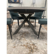 The Rimini Round Extending Table with a gray top and black crossed metal legs, paired with four green quilted-back chairs, creates a stylish, space-saving setup atop a beige and gray patterned rug.
