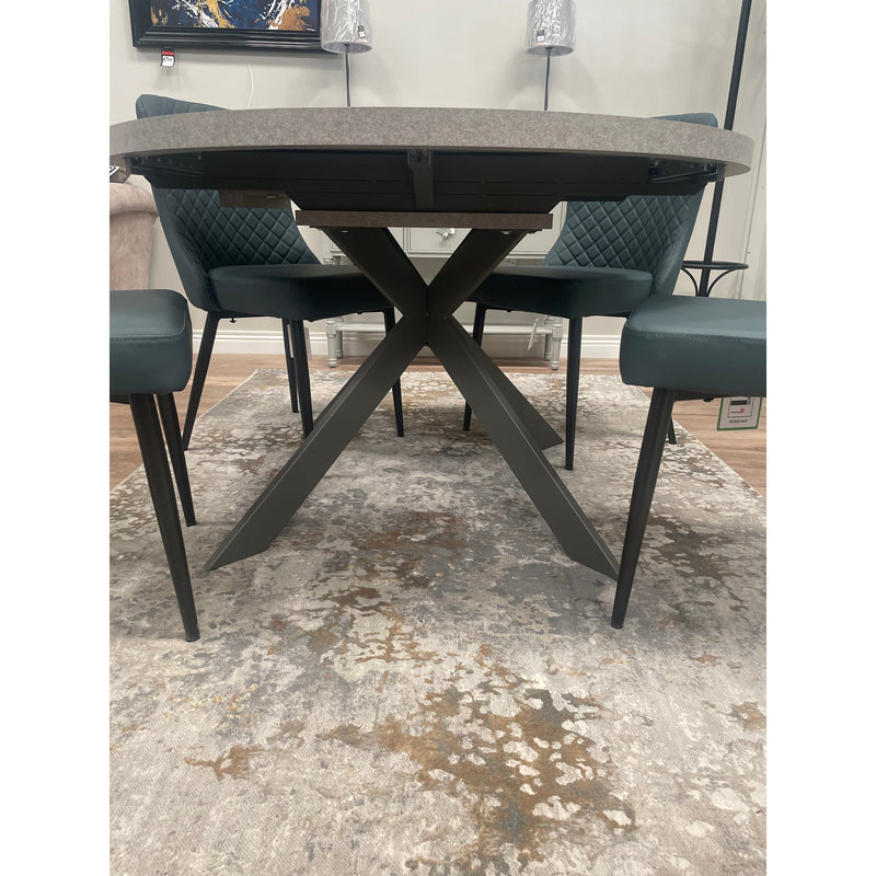 The Rimini Round Extending Table with a gray top and black crossed metal legs, paired with four green quilted-back chairs, creates a stylish, space-saving setup atop a beige and gray patterned rug.