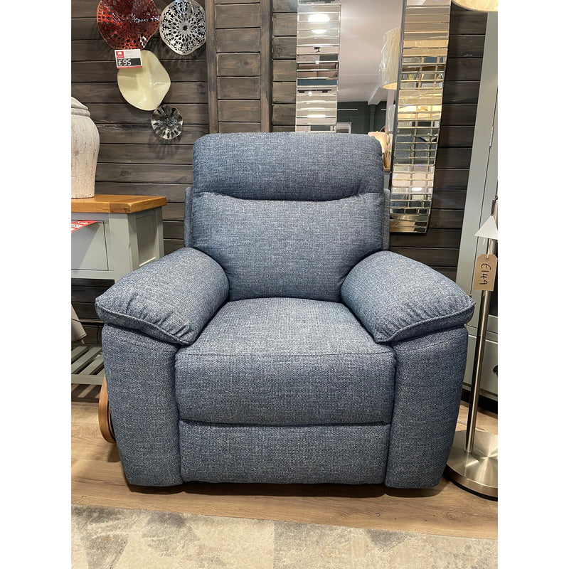 The Vera Recliner Sofa, upholstered in blue with padded armrests and backrest, is displayed on a wooden floor in a showroom among cozy living room furniture and stylish home decor items.