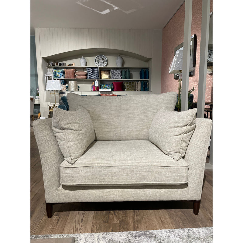 The Layla beige loveseat features Aqua Clean fabric and two matching cushions, set on a wooden floor. Behind it, shelves display vases, books, and décor. Elegant living room furniture accents the space with a pink feature wall on the right.