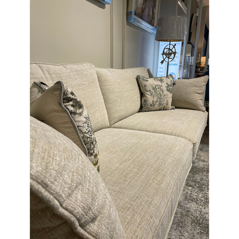 The Layla made-to-measure beige couch with matching patterned throw pillows sits against a neutral wall, beside a floor lamp and grey textured rug. Framed art hangs above this elegant piece of living room furniture.