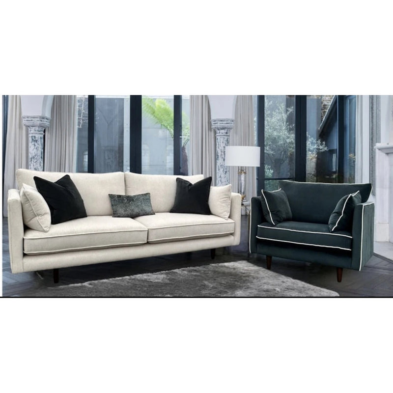A modern living room features the Hugo contemporary sofa in light beige aquaclean fabric and a dark green armchair, both with matching cushions. A gray rug lies on the floor, framed by large windows and decorative pillars.