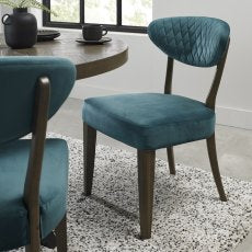 The Ellipse Fumed Oak Upholstered Chair with a teal quilted backrest and dark wooden legs sits by a rustic oak table in a modern dining room featuring a light patterned rug and a large window.