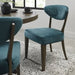 The Ellipse Fumed Oak Upholstered Chair with a teal quilted backrest and dark wooden legs sits by a rustic oak table in a modern dining room featuring a light patterned rug and a large window.