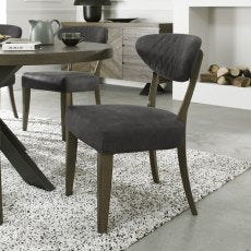 The Ellipse Fumed Oak Upholstered Chair, with dark gray fabric and wooden legs, sits on a white and gray speckled rug beside a rustic oak dining table; a cabinet and stacked firewood appear in the background.