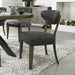 The Ellipse Fumed Oak Upholstered Chair, with dark gray fabric and wooden legs, sits on a white and gray speckled rug beside a rustic oak dining table; a cabinet and stacked firewood appear in the background.