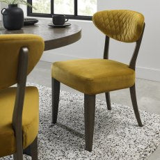 The Ellipse Fumed Oak Upholstered Chair with a quilted backrest and dark wooden legs sits beside a rustic oak dining table in a modern, light-filled room featuring a speckled area rug.