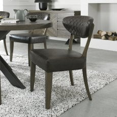 The Ellipse Fumed Oak Upholstered Chair with a dark cushioned seat and backrest sits beside a rustic oak dining table on a speckled rug, with a sideboard, decorative accents, and stacked-firewood fireplace in the background.
