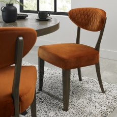 The Ellipse Fumed Oak Upholstered Chair, featuring burnt orange velvet, dark wooden legs, and a quilted backrest, stands beside a rustic oak dining table on a textured rug in a bright, airy room.