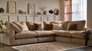 The Eden Corner Group, a cozy beige sectional with deep fiber-filled seats and assorted throw pillows, rests on a patterned rug in a warmly lit living room. Minimalist art, vases, and dried plants adorn a shelf above wainscoting.