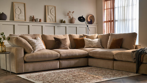 The Eden Corner Group, a cozy beige sectional with deep fiber-filled seats and assorted throw pillows, rests on a patterned rug in a warmly lit living room. Minimalist art, vases, and dried plants adorn a shelf above wainscoting.