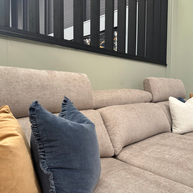 The Nomi Sliding Seat Chaise, a beige fabric customizable sofa with adjustable headrests, is adorned with blue, mustard yellow, and white pillows. It sits against a light green wall featuring black vertical panels above.