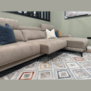 The Nomi Sliding Seat Chaise in beige, topped with blue, white, and brown pillows, rests on a diamond-patterned rug in a living room with green walls and a wall-mounted painting.