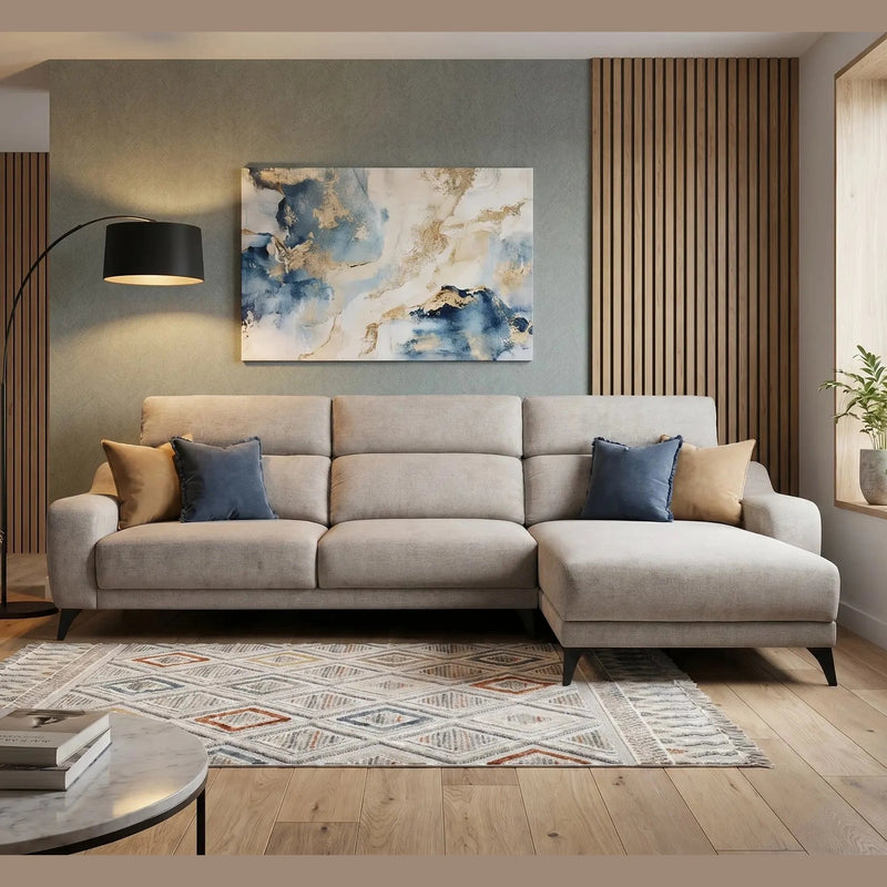 A modern living room featuring the Nomi Sliding Seat Chaise in light beige, blue and tan pillows, a geometric rug, floor lamp, abstract wall art, and a large window with potted plants bathing the space in natural light.