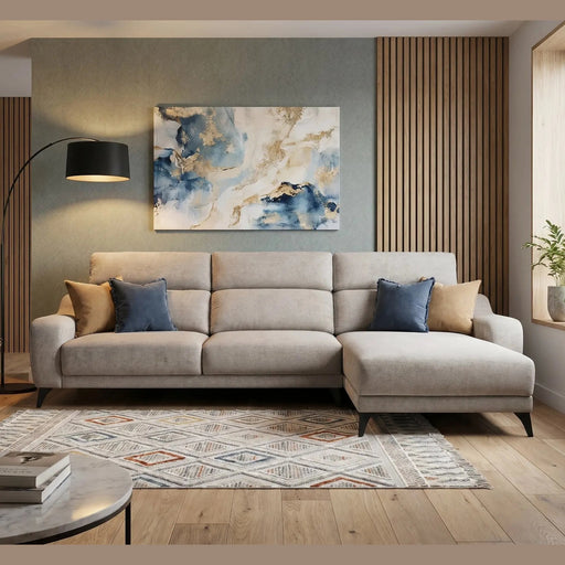 A modern living room featuring the Nomi Sliding Seat Chaise in light beige, blue and tan pillows, a geometric rug, floor lamp, abstract wall art, and a large window with potted plants bathing the space in natural light.