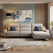 A modern living room featuring the Nomi Sliding Seat Chaise in light beige, blue and tan pillows, a geometric rug, floor lamp, abstract wall art, and a large window with potted plants bathing the space in natural light.