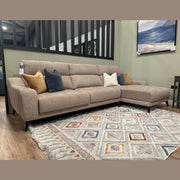 The Nomi Sliding Seat Chaise, a modern beige customizable sofa with five blue, white, and gold accent pillows, sits on a patterned rug in a stylish living room featuring light green walls and a framed landscape painting.
