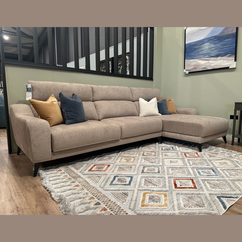 The Nomi Sliding Seat Chaise, a modern beige customizable sofa with five blue, white, and gold accent pillows, sits on a patterned rug in a stylish living room featuring light green walls and a framed landscape painting.