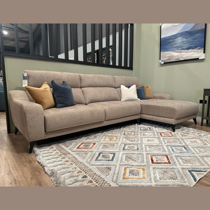 The Nomi Sliding Seat Chaise, a modern beige customizable sofa with five blue, white, and gold accent pillows, sits on a patterned rug in a stylish living room featuring light green walls and a framed landscape painting.