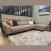 The Nomi Sliding Seat Chaise, a modern beige customizable sofa with five blue, white, and gold accent pillows, sits on a patterned rug in a stylish living room featuring light green walls and a framed landscape painting.