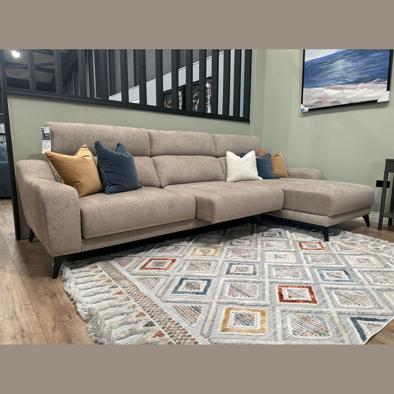 The Nomi Sliding Seat Chaise, a modern beige customizable sofa, features assorted blue, white, and tan pillows and sits on a patterned rug in a stylish living room with wood flooring and framed artwork.