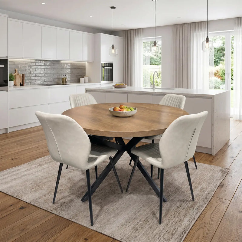A modern kitchen with white cabinets, a large island, and wooden floors features the Panama Table with 4 Lyra Chairs on a beige rug. A fruit bowl decorates the table as natural light streams in through large windows.