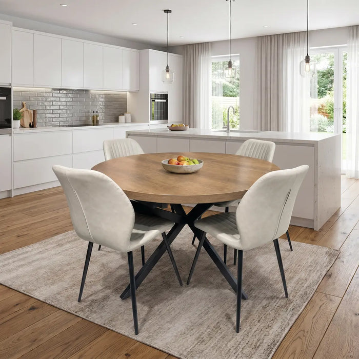 A modern kitchen with white cabinets, a large island, and wooden floors features the Panama Table with 4 Lyra Chairs on a beige rug. A fruit bowl decorates the table as natural light streams in through large windows.