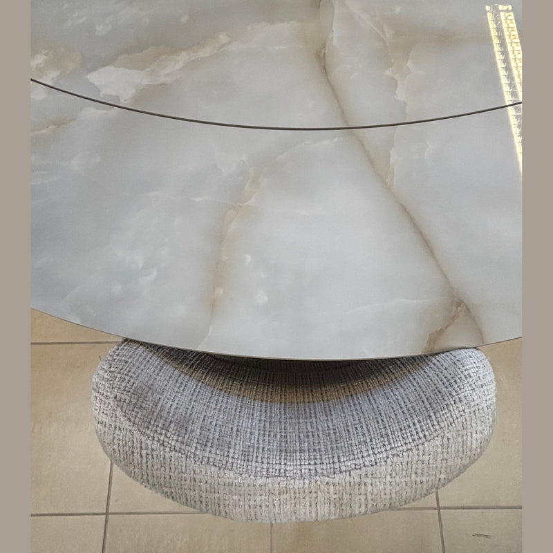 The Positano Swivel Dining Table showcases a round, cloudy-patterned ceramic top with a seam and black metal legs, accompanied by a circular textured gray upholstered stool, both set on a tiled floor.