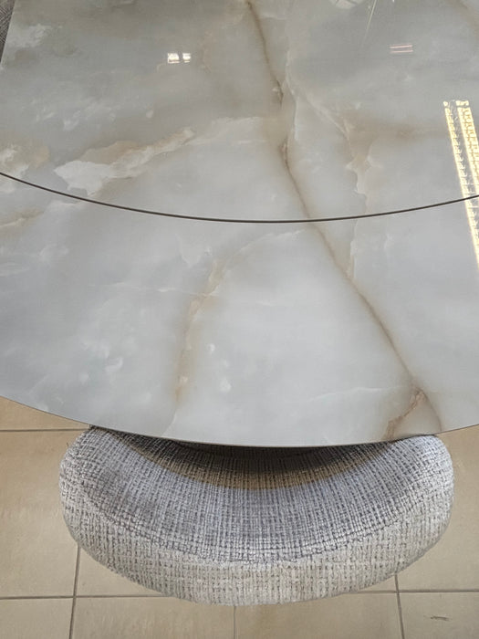 The Positano Swivel Dining Table showcases a round, cloudy-patterned ceramic top with a seam and black metal legs, accompanied by a circular textured gray upholstered stool, both set on a tiled floor.