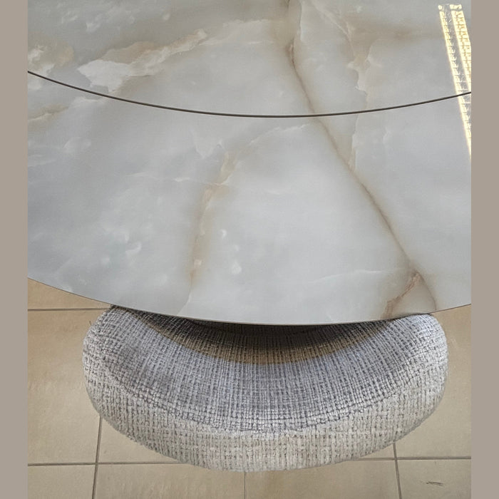 The Positano Swivel Dining Table showcases a round, cloudy-patterned ceramic top with a seam and black metal legs, accompanied by a circular textured gray upholstered stool, both set on a tiled floor.