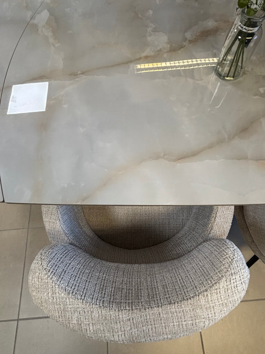 A top-down view of the Positano Swivel Dining Table with a ceramic top and black metal legs, a glass vase with green stems in the top right corner, a textured cushioned chair tucked in, and tiled flooring below.