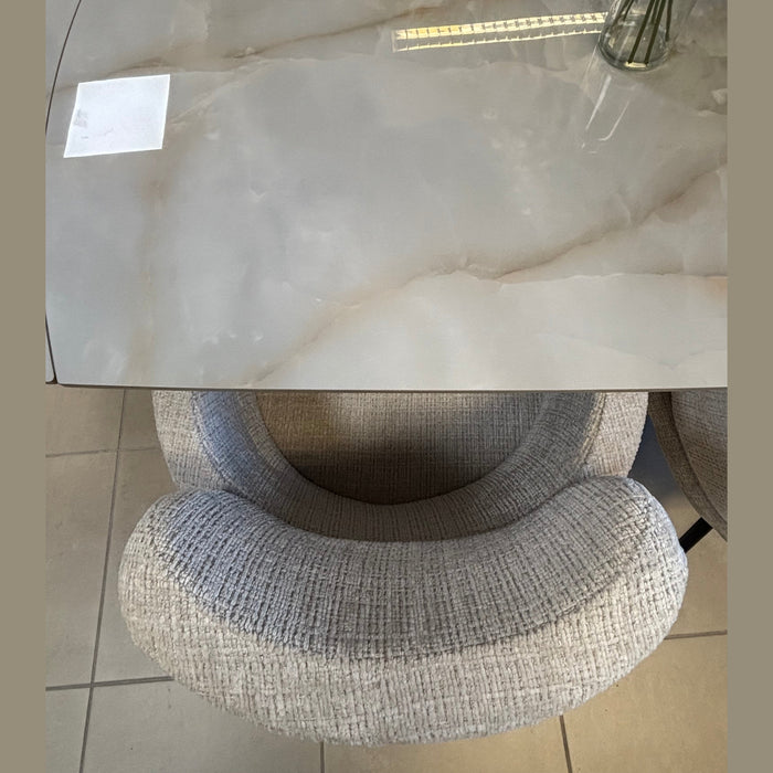 A top-down view of the Positano Swivel Dining Table with a ceramic top and black metal legs, a glass vase with green stems in the top right corner, a textured cushioned chair tucked in, and tiled flooring below.