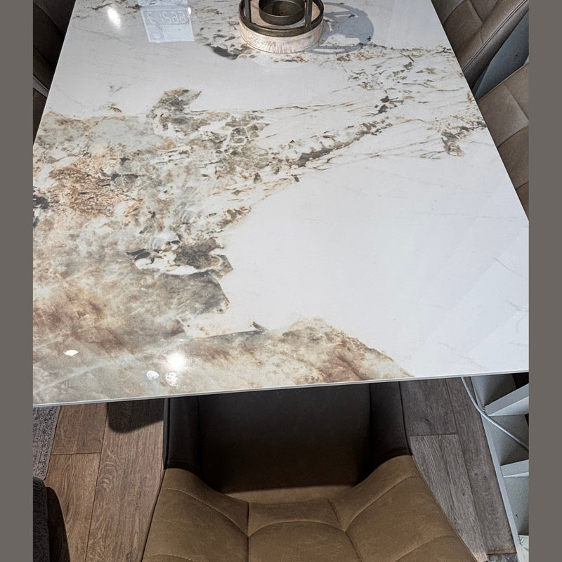 The Sorrento Dining Table, featuring a glossy marble effect with brown and beige patterns, is paired with beige upholstered seating and rests on a wooden floor, complete with a stylish round centerpiece.