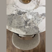 The Fusion Dining Table, featuring a gray and white marble-inspired top, rests on wooden flooring. A textured light gray upholstered chair sits beside it, while a gold accent decorates the tabletop.
