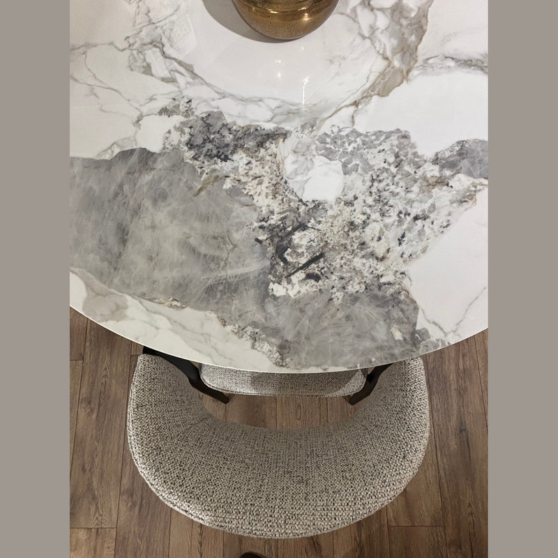 The Fusion Dining Table, featuring a gray and white marble-inspired top, rests on wooden flooring. A textured light gray upholstered chair sits beside it, while a gold accent decorates the tabletop.
