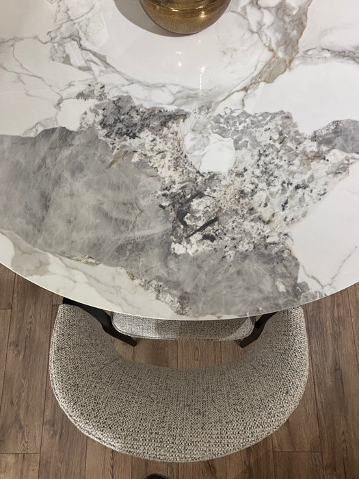 The Fusion Dining Table, featuring a gray and white marble-inspired top, rests on wooden flooring. A textured light gray upholstered chair sits beside it, while a gold accent decorates the tabletop.