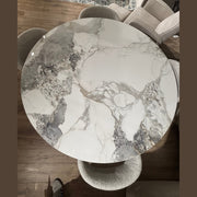 The Fusion Dining Table, featuring a marble-effect top with white, gray, and beige veining, is paired with four upholstered chairs on a wooden floor, partial patterned rug, and other furniture visible in the background.