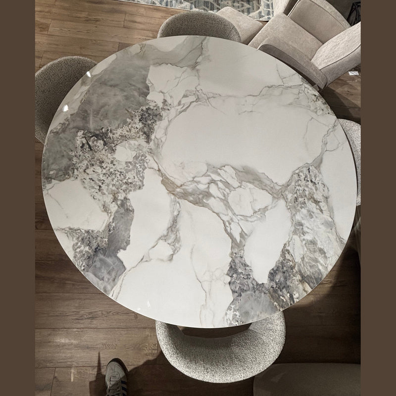 The Fusion Dining Table, featuring a marble-effect top with white, gray, and beige veining, is paired with four upholstered chairs on a wooden floor, partial patterned rug, and other furniture visible in the background.