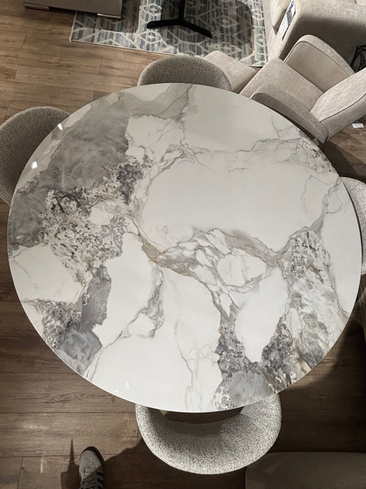 The Fusion Dining Table, featuring a marble-effect top with white, gray, and beige veining, is paired with four upholstered chairs on a wooden floor, partial patterned rug, and other furniture visible in the background.