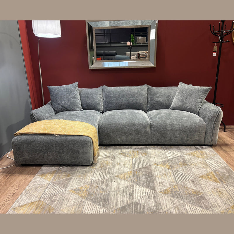 The Izzy Chaise Corner in modern grey, featuring plush cushions and a yellow blanket on the chaise section, sits on a patterned rug. A large mirror is mounted on the deep red wall behind the sofa.