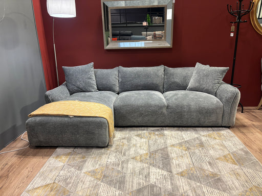 The Izzy Chaise Corner in modern grey, featuring plush cushions and a yellow blanket on the chaise section, sits on a patterned rug. A large mirror is mounted on the deep red wall behind the sofa.