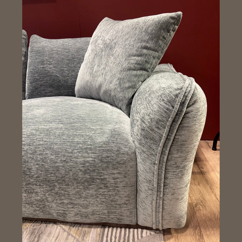 A close-up of the Izzy Chaise Corner, a modern grey sofa with a large cushion, set against a red wall and placed on a light wood floor with a patterned area rug.