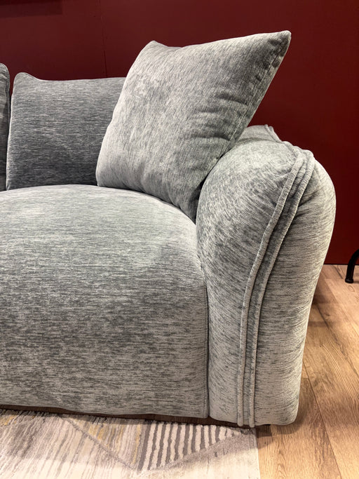 A close-up of the Izzy Chaise Corner, a modern grey sofa with a large cushion, set against a red wall and placed on a light wood floor with a patterned area rug.