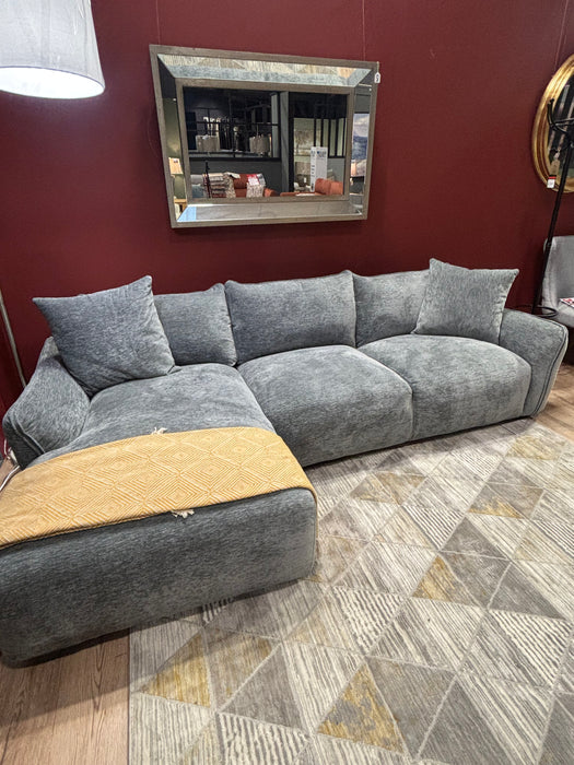 A modern grey Izzy Chaise Corner sofa with large cushions sits on a geometric-patterned rug. A mustard yellow throw is draped over the chaise, and a rectangular mirror hangs on a dark red wall behind.