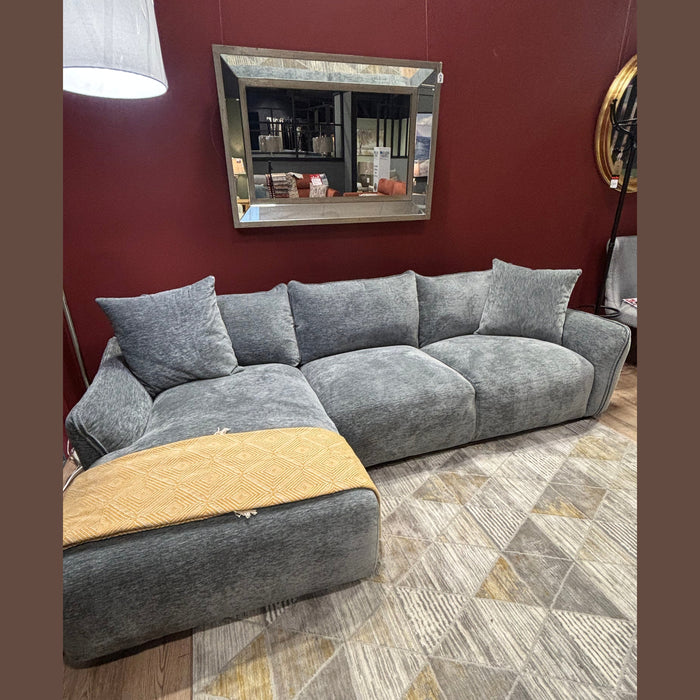 A modern grey Izzy Chaise Corner sofa with large cushions sits on a geometric-patterned rug. A mustard yellow throw is draped over the chaise, and a rectangular mirror hangs on a dark red wall behind.