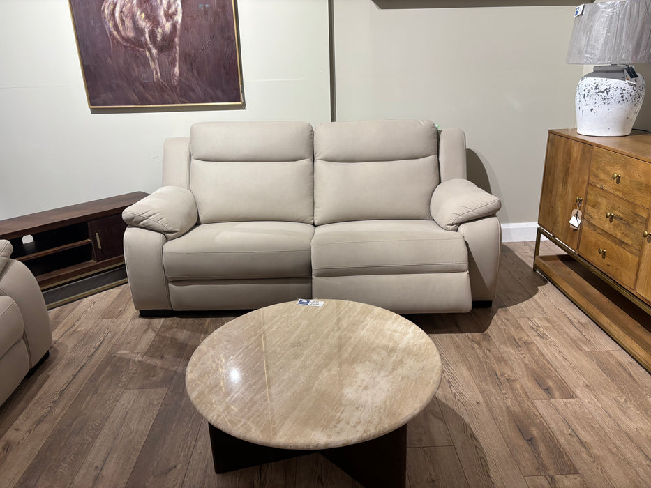 The Lorenzo Electric Recliner in modern beige sits against a light wall with artwork above, facing a round wooden coffee table. To the right, a wooden cabinet displays a white vase and lamp.