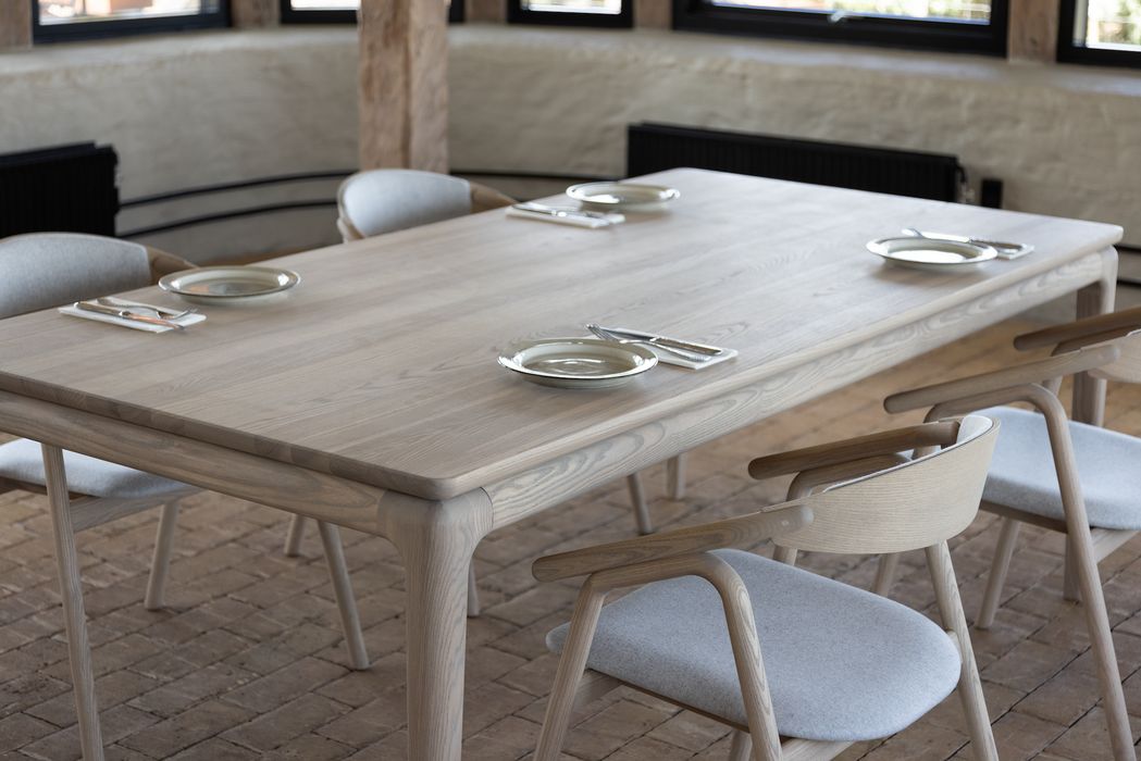 The Floating Dining Table, custom crafted from ash wood with six matching chairs, is set and ready. Brick floors, wooden beams, and wall windows enhance the inviting atmosphere of the dining space.