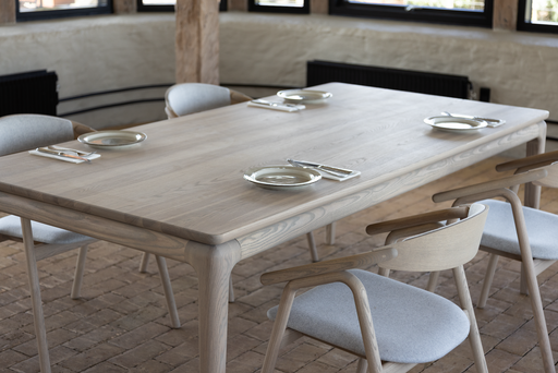 The Floating Dining Table, custom crafted from ash wood with six matching chairs, is set and ready. Brick floors, wooden beams, and wall windows enhance the inviting atmosphere of the dining space.
